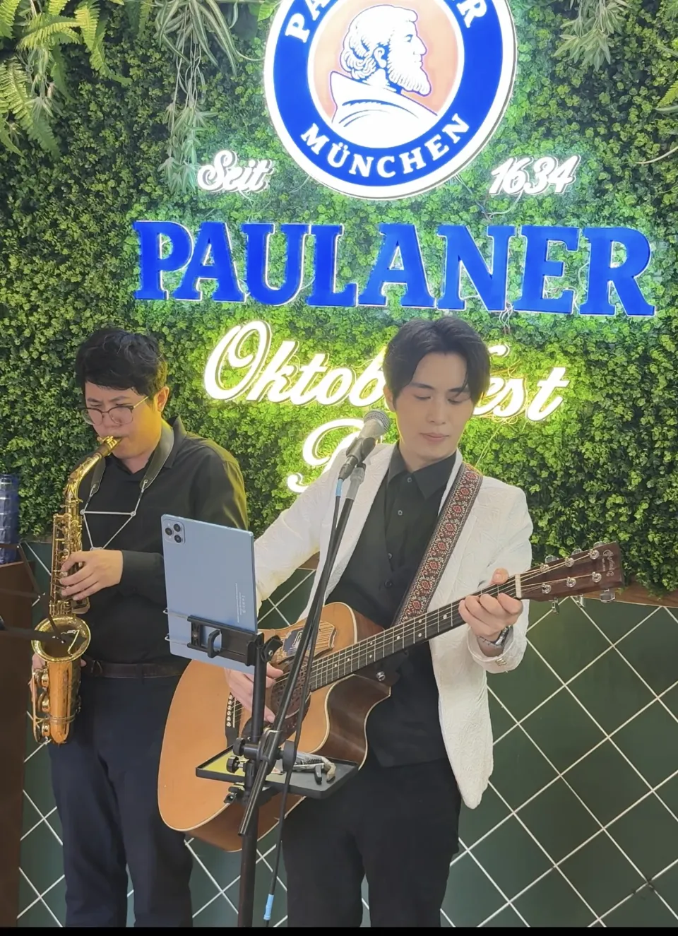 品牌活動實景：綠色植物牆前，一位樂手吹奏 Saxophone，另一位穿白色外套的樂手彈木結他並唱歌，面前是一支咪高峰與夾著平板電腦的譜架
